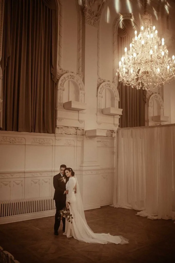 Couple standing beneath a chandelier in the ornate hall at Maison Principale