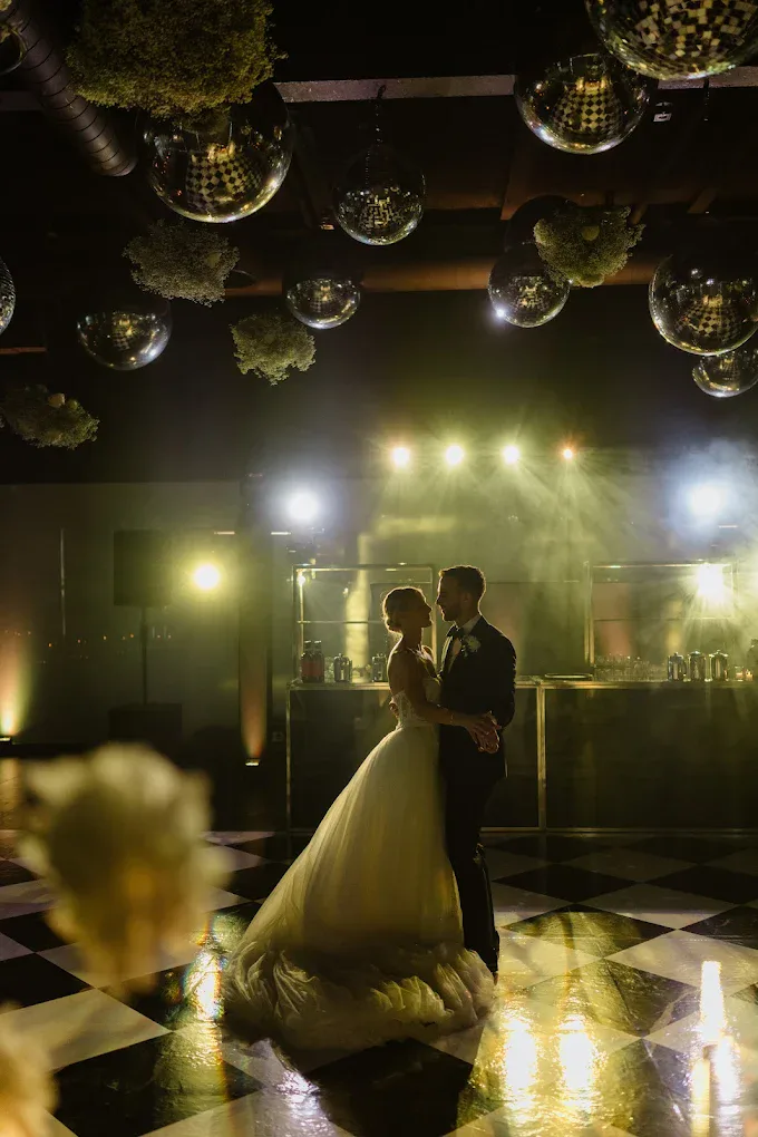 Couple slow dancing on a checkered floor under mirror balls, stage lights glowing behind them